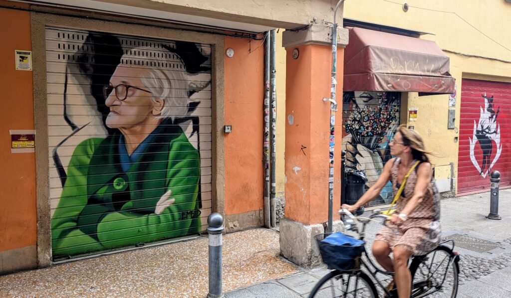 a piece of street art in bologna italy depicting an old grandma looking very stern, with a lady on bike cycling past it and looking.