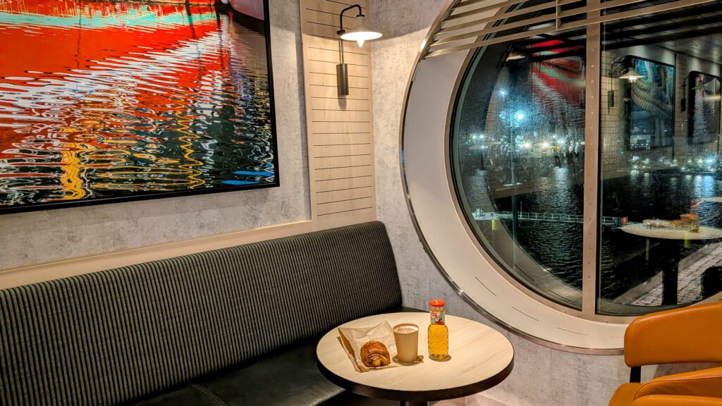 a pain au chocolat, bottle of apple juice, and a coffee on a table with some seating and a porthole in the background, with portsmouth port by night out of the window.